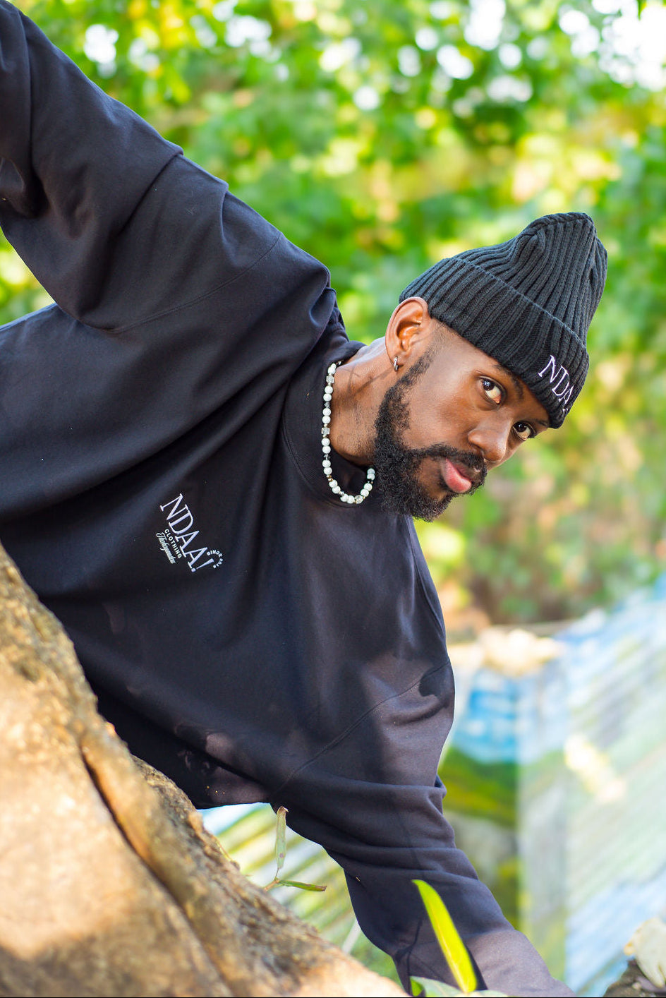 A man wearing a black ribbed beanie with white 'NOLA' lettering and a black sweatshirt with white 'INDLAG' text, posing outdoors on a rock with a green bokeh background.